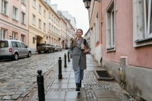 A smiling woman walks down a charming cobblestone street holding books, showcasing urban lifestyle.